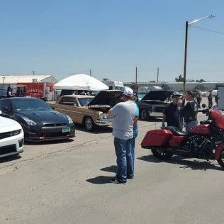 A group of people are standing in a parking lot looking at cars and a motorcycle.