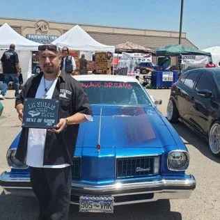 A man is standing in front of a blue car holding a sign.