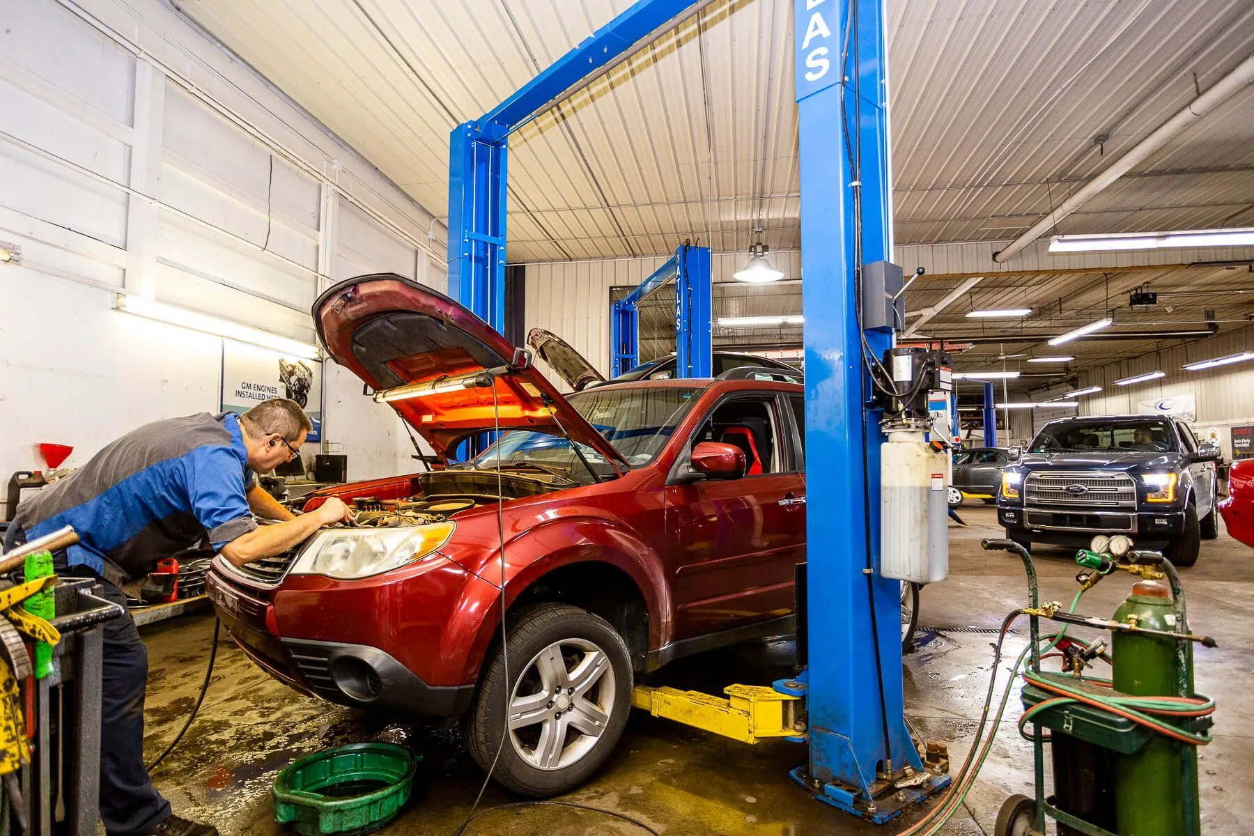 Technician working under the open hood of a red SUV on a lift in an auto repair shop