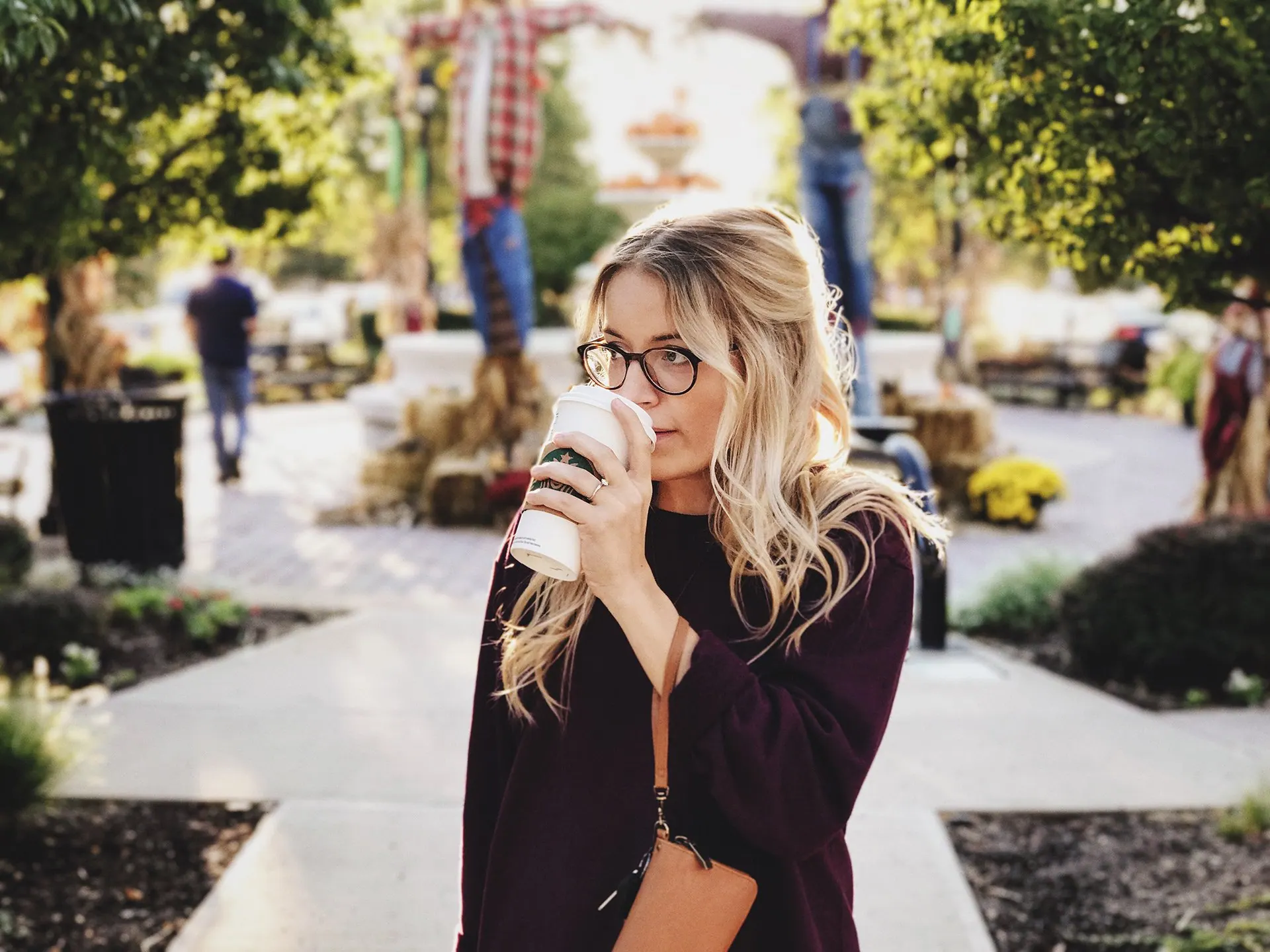 Woman in glasses drinking coffee on a walkway with autumn decorations